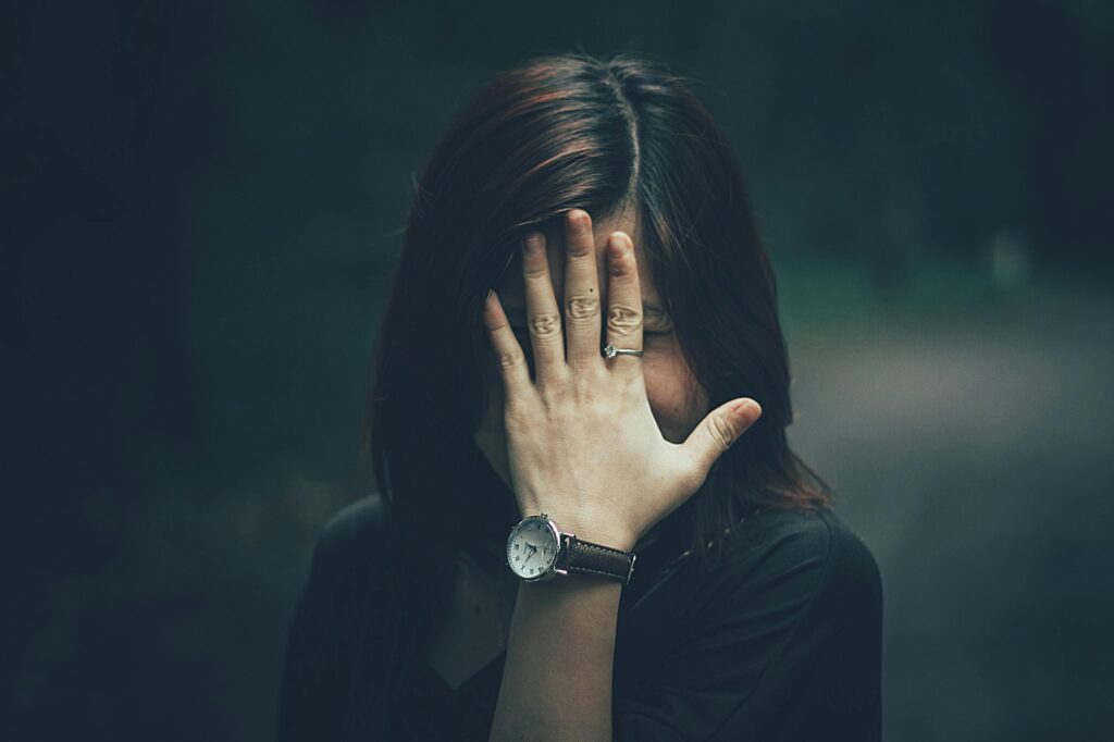 A woman with a wristwatch hides her face with her hand in a moody outdoor setting.
