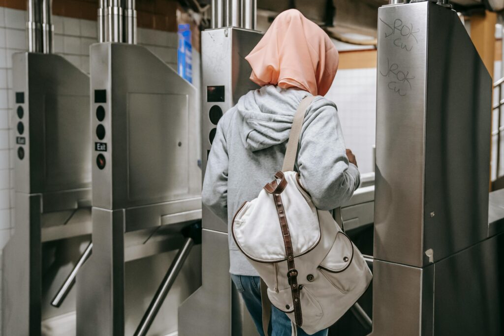 Back view of unrecognizable Muslim woman in casual clothes with backpack and hijab walking through turnstile in metro